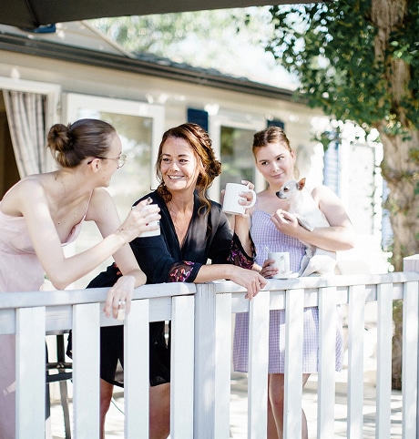 Moment de complicité entre femmes sur une terrasse Trois femmes souriantes discutent sur une terrasse, tenant des tasses et un petit chien blanc.